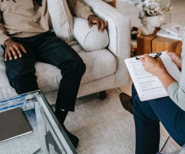 From above crop African American male in casual clothes sitting on comfy couch and answering professional female consultant questions in modern psychotherapy office